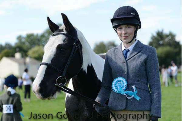 woman dressed in show gear with a rosette pinned to her jacket, stood next to a black and white cob