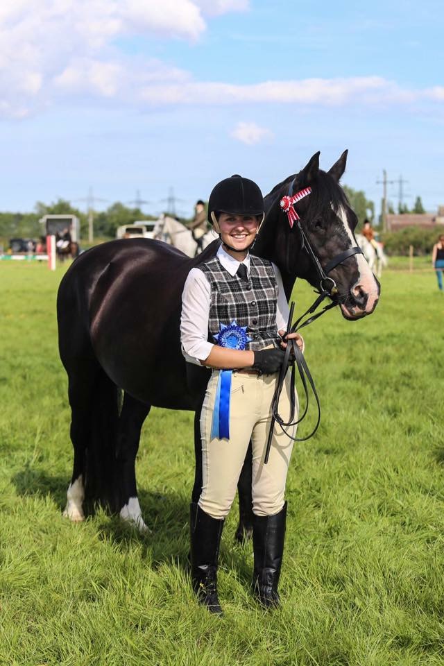 woman dressed in show gear standing with her black cob, both wearing rosettes