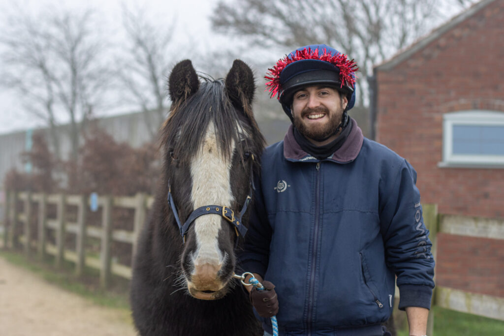 Handler stood holding the lead rope of a black cob, he is wearing tinsel on his hat