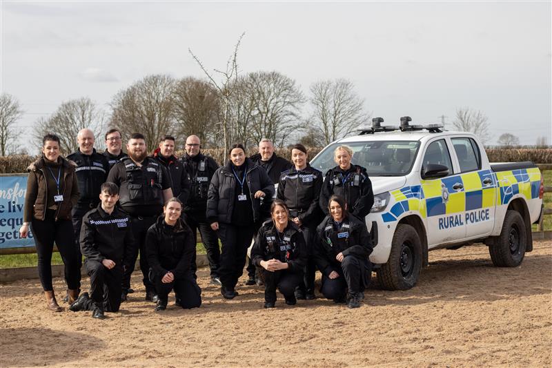 Group of Nottinghamshire Police rural crime officers and Bransby Horses staff standing and kneeling together in front of a marked Rural Police vehicle, outdoors on a sandy area with trees and a hedgerow in the background.