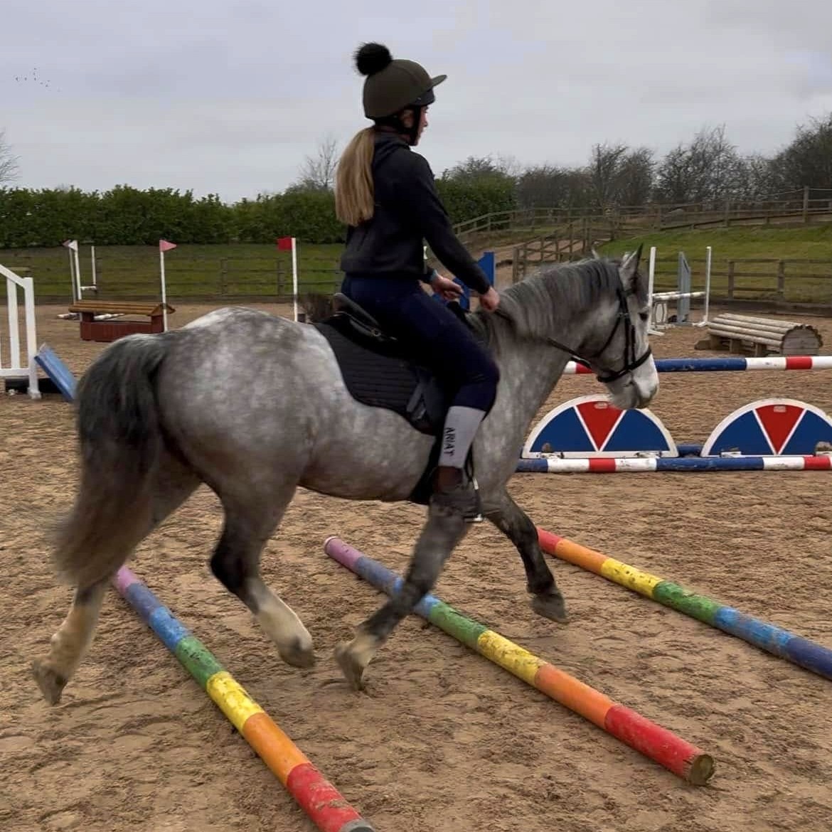 a woman riding a grey dappled pony over some trotting poles
