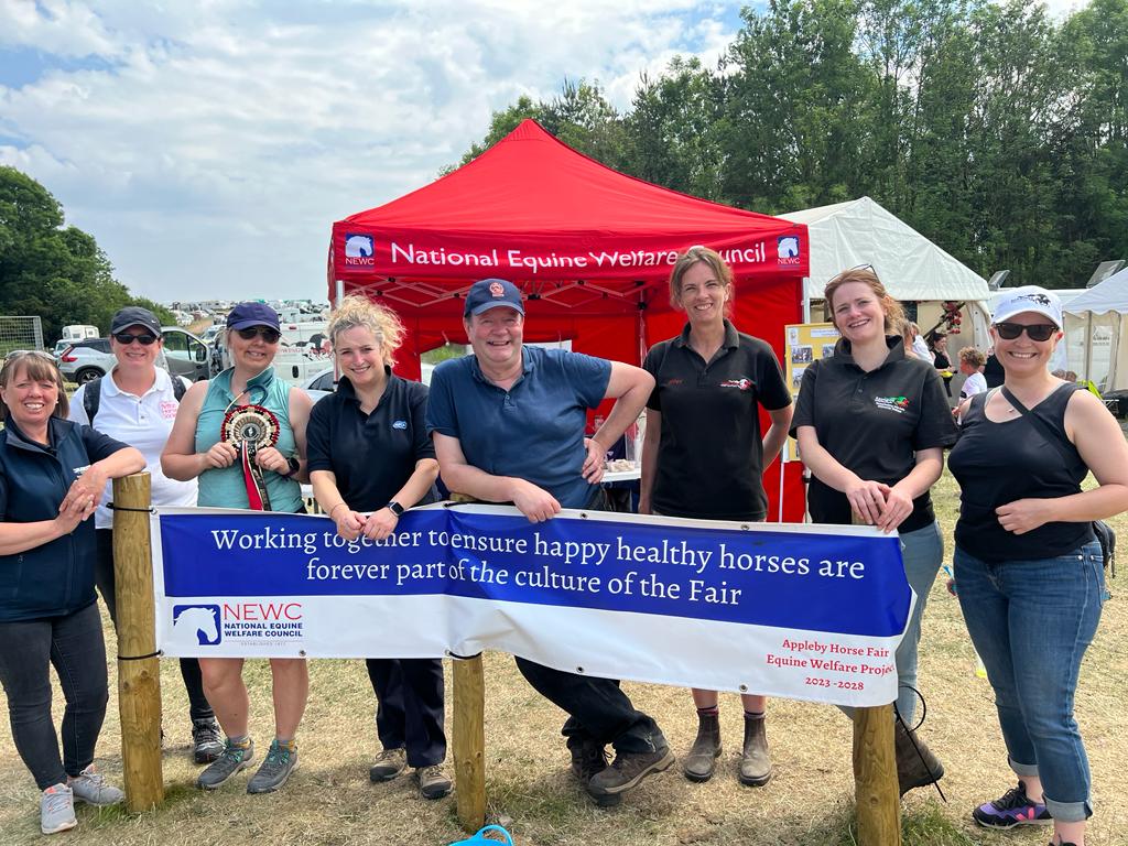 A group of charity staff stand together at Appleby Fair in front of a red National Equine Welfare Council (NEWC) tent, holding a banner about working together to ensure happy, healthy horses at the Fair.